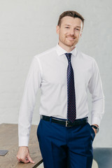 Indoor shot of handsome unshaven male entrepreneur wears formal shirt, tie and trousers, keeps hand...