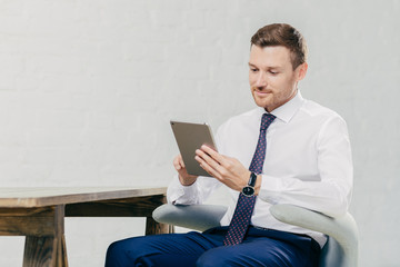 Prosperous handsome young businessman watches webinar online on modern tablet computer, dressed in formal clothes, sits at chair indoor. Young attractive male financier reads news on web page online