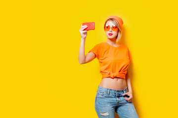 Young style girl with orange glasses make a selfie with her phone on yellow background. Clothes in 1980s style
