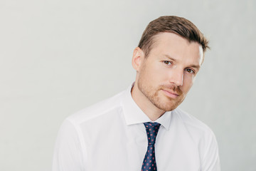 Confident male looks seriously at camera, has dark eyes and stubble, dressed in elegant white shirt with tie, isolated over white background. Man director thinks about how reach success in life