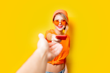 Young style girl with orange glasses give to you her phone on yellow background. Clothes in 1980s style