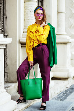Outtdoor full body portrait of young beautiful fashionable woman wearing stylish headband, yellow blouse, violet trousers, sunglasses, hodilng green leather bag, posing in street of european city.