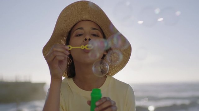close up portrait of happy young hispanic woman blowing bubbles enjoying relaxed summer vacation lifestyle on beautiful sunny beach wearing hat