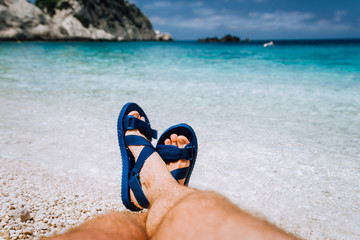 Fototapeta premium Młody mężczyzna skrzyżował stopy w niebieskich sandałach flip-flop opalając się na plaży