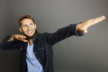 Young bearded cheerful man in casual denim jacket doing dabbing posture on gray background smiling away