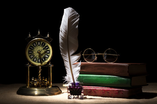 Vintage still life with retro glasses on old books near inkstand, feather, old clock against black background under beam of light. Dramatic background. Literature concept.