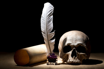 Drama or theater concept. Old inkstand with feather near scroll with skull against black background. Dramatic light