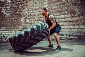 Muscular bearded tattooed fitness man moving large tire in street gym. Concept lifting, workout...