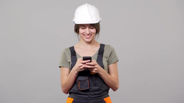 Portrait of sociable woman builder in helmet and jumpsuit smiling and typing text message on smartphone, isolated over gray background