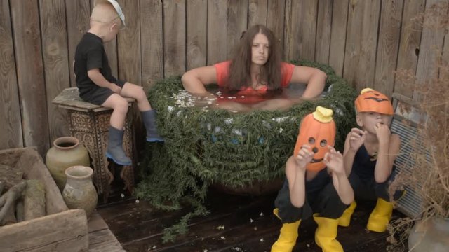 Young witch flew on broom in secluded place to plunge into cauldron with decoction of rejuvenating herbs on eve of Halloween. Happy Children dressed in pumpkin masks play with witch and tease her