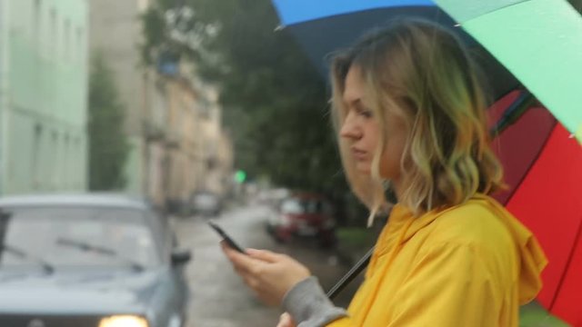 calm serious business woman using smart phone mobile while standing outside on the street in rainy cloudy day holding an umbrella using taxi application online app communication internet notification