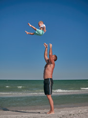 Obraz premium Dad and Baby playing on seaside in summertime. Happy family and happy childhood concept. Little boy having fun with Daddy on beach. Dad throwing and catching up a kid into air. Summer rest concept