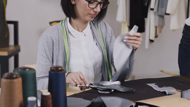 Close-up of a dressmaker that sits at the table and cuts pattern from gray material with scissors for sewing clothes in a small stylish atelier. Young woman tailor working with scissors at the studio