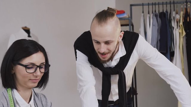 Close-up of two tailor caucasian fashion designers working with gray material using pattern sitting at the beautiful office with different tailoring tools and clothes. Tailor and dressmaker create a