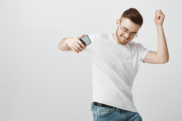 Joyful pleased and carefree exciting handsome unshaven guy in glasses holding black smartphone...
