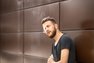 A young handsome white bearded man in black t-shirt smokes a cigarette in the street in the spring. Close up.
