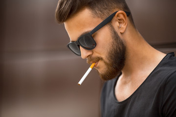 A young handsome white bearded man in black t-shirt smokes a cigarette in the street in the spring....