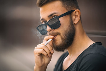 A young handsome white bearded man in black t-shirt smokes a cigarette in the street in the spring. Close up.
