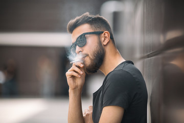 A young handsome white bearded man in black t-shirt smokes a cigarette in the street in the spring....