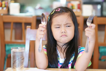 Asian children hungry or kid girl holding spoon with fork and unhappy or angry and touchy frown for long time wait eat delicious food or anorexia and bored food for breakfast in morning at restaurant