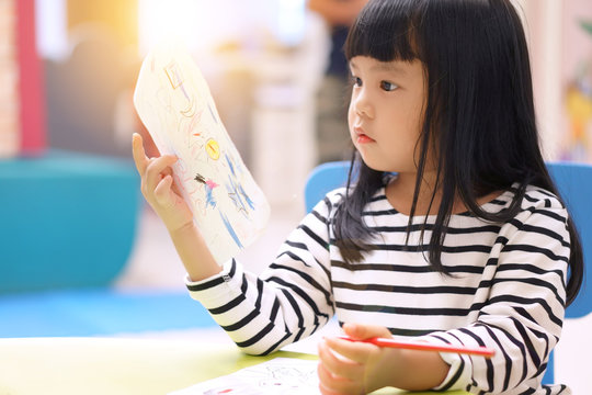 Asian children cute or kid girl and student learning for study coloring or drawing paint on paper and colorful table with chair at nursery or pre school and kindergarten on soft and sunlight flare