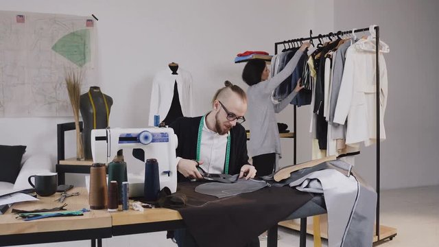 Tailor or fashion designer sits on the workplace at studio and cutting gray fabric using large scissors as he follows the chalk markings of the pattern using pattern. In the background, the girl looks