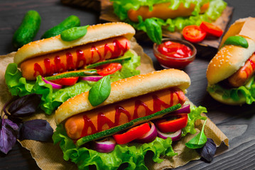  Homemade sandwiches sausages in buns (hot dogs). Ingredients for hot dogs onions, fresh lettuce, tomatoes, cucumbers. ketchup, basil. Dark wooden background.