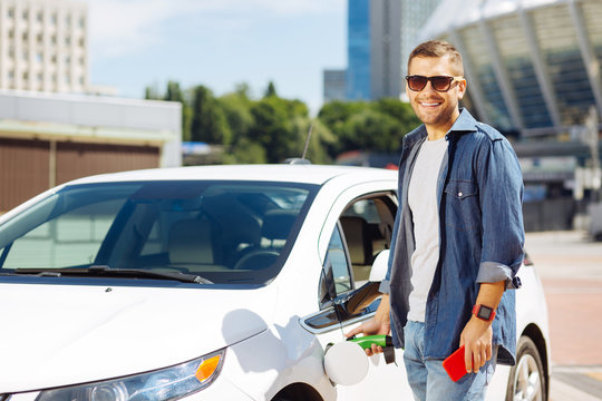 Positive mood. Cheerful handsome man smiling to you while fuelling his car