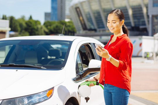 Modern age. Confident young woman looking at her smartphone while holding a fuel nozzle