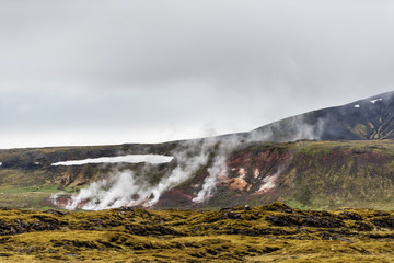 Landscape view of Iceland mountains with steam from hot springs geysers during cloudy, overcast day, nobody in distant, cliff and snow during spring, summer