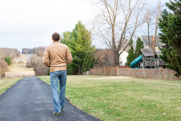 Back of young man walking on Sugarland Run Stream Valley Trail hike in Herndon, Northern Virginia, Fairfax county residential neighborhood in winter, spring, paved path road, houses
