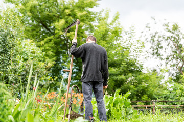 Back of young man farmer in garden standing with sickle scythe tool in green summer in Ukraine by well faucet