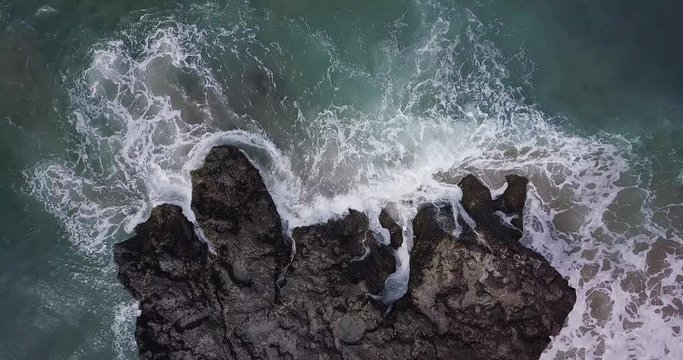 Aerial top view waves break on dark rocks near beach. Sea waves on the dangerous stones aerial view drone 4k shot. Bird's eye view of ocean waves crashing against an empty beach from above.