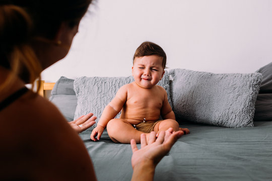 Annoyed baby crying sitting on the bed in the room wearing shorts, mom is in the frame of the image animating him, frustration concept