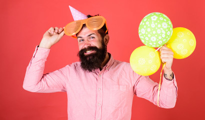 Hipster peeking out of giant sunglasses. Guy in party hat with air balloons celebrates. Celebration concept. Man with beard and mustache on happy face holds bunch of air balloons, red background