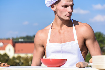 Chef cook with nude sexy muscular torso. Man on confident face wears cooking hat and apron, skyline on background. Cook or chef with muscular shoulders and chest looks attractive. Masculinity concept
