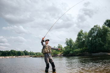 Fisherman stands in shallow and holding fly rod up in one hand and part of spoon in the other one. Man is looking up. He is calm and concentrated.