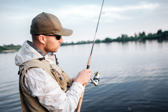 A picture of man in cap, sunglasses and vest standing in shallow and looking at fly rod. He is using non-inertial reel. Guy is concentrated.