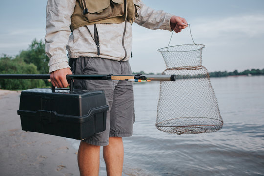 A picture of man holding fishing net and plastic black box in hands. Also he has fly rold in the right one. Guy is standing at the watr shore.