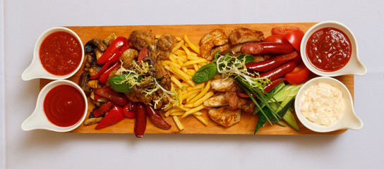 Wooden dish with vegetables, mushrooms, sausage and fried meat. Four various sauces in elegant dishes and greenery. On a white table-cloth. A photo is done from above.
