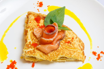 Pancakes with red caviar, slices of red fish, greenery and sauce on a white dish