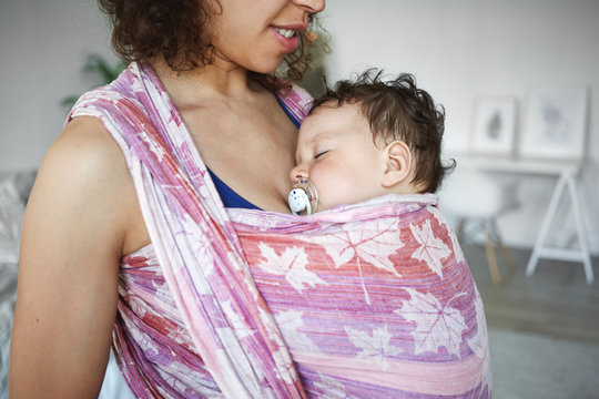 Close up shot of adorable cute newborn kid sucking on pacifier while sleeping peacefully in baby carrier at his mother's breast. Cropped image of young dark skinned mother carrying son in wrap