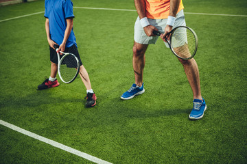 Obraz premium Man and child playing tennis while situating on green field for game outdoor. They wearing modern trainers