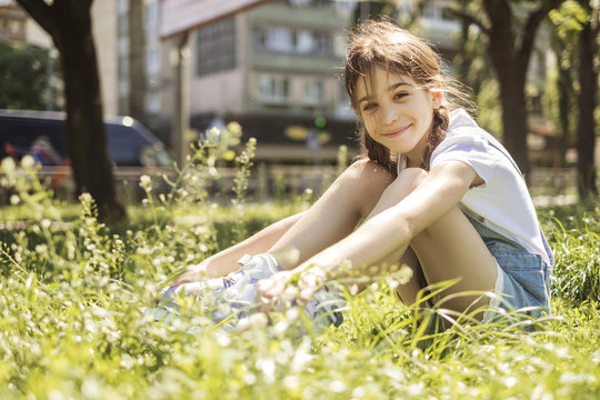 Cute little 10 years old girl in casual outfit playing at park in warm summer day. She sitting in grass, smiling. Copy space