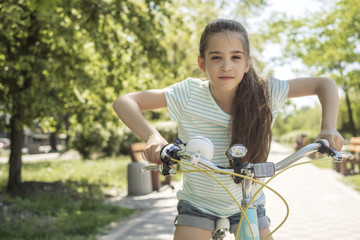 Obraz premium Cute little 10 years old girl in casual outfit playing at park in warm summer day. She having fun riding a bicycle