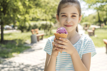 Cute little 10 years old girl in casual outfit playing at park in warm summer day. She have fun licking an ice cream in  a waffle cup. Copy space