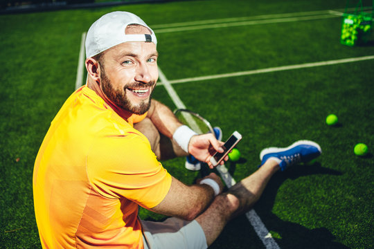Portrait of cheerful bearded man having leisure with mobile after good training. He sitting on court