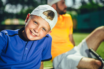 Portrait of happy boy with attractive smile wearing cap. He looking at camera. Calm man resting behind him after playing tennis