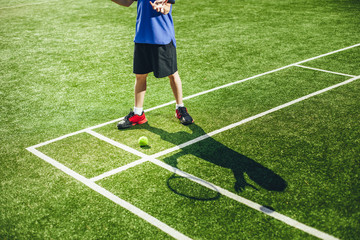 Close up child feet and his silhouette situating on field. He throwing ball and keeping racket