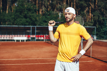 Portrait of smiling young bearded playing in tennis while situating on court outdoor during sunny day. He keeping tennis racquet on shoulder and wearing cap on head